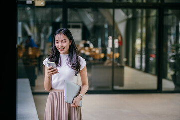 Poster - Young asian businesswoman wearing a white t-shirt and beige skirt, walking confidently near an office building while holding a laptop and using her smartphone for communication