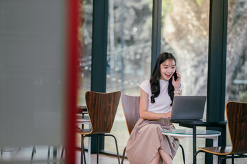 Poster - Smiling young asian freelancer woman is sitting at a table in a cafe, talking on her phone and working on her laptop, enjoying the natural light and modern ambiance