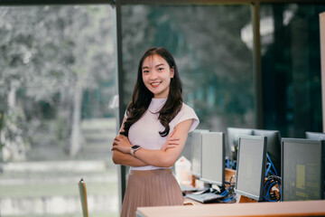 Young professional woman is standing in a computer lab with her arms crossed, smiling confidently, showcasing her expertise and leadership in the tech industry