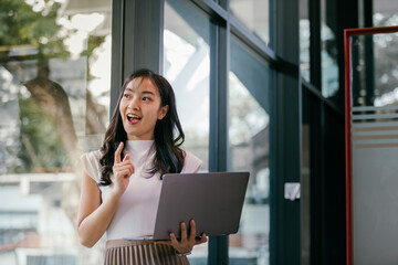 Poster - Asian businesswoman is holding a laptop and having a new idea while working in a modern office, she looks excited and points her finger up