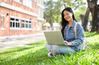 © bongkarn - A smiling Asian female college student sitting on the grass in her university park, using her laptop