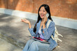 © bongkarn - A positive Asian female student enjoys a phone conversation while sitting on the edge of a footpath.