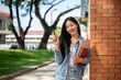 © bongkarn - A female college student stands leaning against a brick building wall, smiling at the camera.