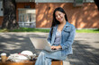 © bongkarn - A positive Asian female college student sits on a bench at her university, using her laptop to work.