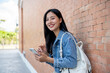 © bongkarn - Smiling female student stands leaning against a brick building wall outside, holding her smartphone.