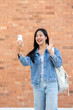 © bongkarn - A cheerful Asian female student talks on a video call through her smartphone while standing outdoors