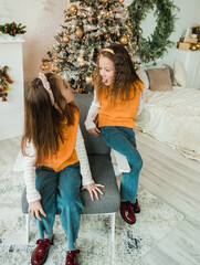 Wall Mural - Two playful sisters, dressed in matching orange sweaters and jeans, share a silly moment by the Christmas tree