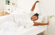 © Studio Romantic - Cheerful African American girl with curly hair lying in bed, waking up in bright sunlit bedroom. Happy smiling black woman stretching after restful night sleep or relaxing nap, feeling refreshed.