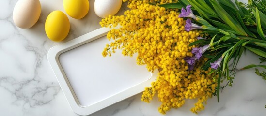 Poster - Spring Floral Arrangement with Yellow Mimosa and Purple Irises Next to an Empty White Frame and Colorful Eggs on Marble Surface Flat Lay