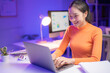 © MINAE - Happy businesswoman using laptop computer typing on keyboard looking at screen working late at night in office with charts showing on desktop screen and desk lamp illuminating workspace