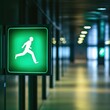 © Chexy139 - A glowing green emergency exit sign shows an individual running, guiding people towards safety in a well-lit, contemporary hallway of a public building