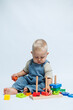 © DSMT - Cute blond boy in dungarees sits on a white background and plays with a wooden sorter. Sorter for the development of fine motor skills of babies. Child plays with educational toys.