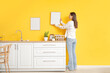 © Pixel-Shot - Young woman hanging blank frame on yellow wall in kitchen