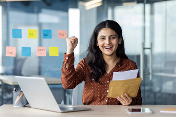 Wall Mural - A cheerful young woman celebrates at her desk, holding an envelope with joy. Her laptop and phone are on the desk, signifying a successful outcome.