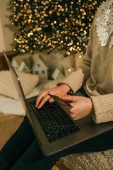 Wall Mural - Woman using a laptop with a credit card in hand, shopping online during the Christmas season, with a glowing festive tree in the background creating a cozy holiday vibe