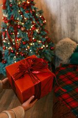 Wall Mural - Person unwrapping a festive gift box with a red ribbon near a beautifully decorated Christmas tree. Cozy holiday atmosphere with pillows, candles, and glowing fairy lights in the background