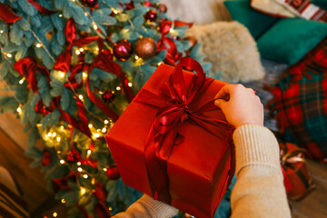 Wall Mural - Person unwrapping a festive gift box with a red ribbon near a beautifully decorated Christmas tree. Cozy holiday atmosphere with pillows, candles, and glowing fairy lights in the background