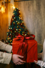 Hands exchanging a festive gift box with a red ribbon under a beautifully decorated Christmas tree. Cozy holiday setting with candles, miniature houses, and a warm atmosphere