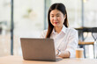 © David - Portrait of a business asian woman sitting in an office with her colleagues in the background. Happy business woman working in a co-working office.