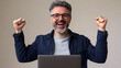 © Vitaliy - A joyful Middle-Eastern man in his 40s celebrates success, raising his fists in excitement while sitting at his laptop in a minimalistic setting.