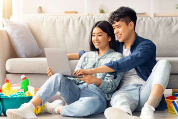 Wall Mural - Relaxed young asian couple want to clean apartment, sitting on floor by couch in living room, smiling and hugging, using modern laptop, looking for house-keeping service online, copy space