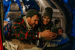 © La Famiglia - Two people drinking tea in a cozy car trunk setup and using a phone together.