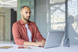 © Liubomir - Confident businessman working diligently on a laptop in a bright, modern office. The professional setting includes essentials like a notebook, pen, and tablet, emphasizing focus and productivity.