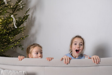 Wall Mural - Two sisters peek over a light gray couch, their expressions full of playful surprise. A decorated Christmas tree is visible in the background.