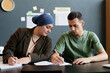 © Seventyfour - Medium shot of adult man explaining task to female Mulsim fellow student writing in exercise book while sitting at desk in English language school classroom