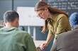 © Seventyfour - Medium shot of young female teacher standing by adult student at desk explaining homework task at end of lesson in English language school, copy space