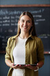 © Seventyfour - Vertical portrait shot of happy female English teacher posing with tablet computer in hands while standing against blackboard in school classroom, copy space