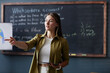 © Seventyfour - Medium shot of female English teacher holding tablet computer addressing student with question, while discussing new grammar rules in group class against blackboard at language school