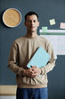 © Seventyfour - Vertical portrait shot of happy mixed raced male student posing with exercise book in hands while standing against blue wall in school classroom, copy space