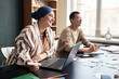 © Seventyfour - Medium shot of cheerful Muslim woman in headscarf conversing with fellow student practicing new vocabulary in speaking club group while using laptop at large desk