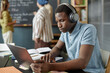 © Seventyfour - Concentrated African American male student in headphones using laptop completing listening task in English class at language school for adult learners, copy space