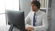 © Krakenimages.com - Young man working in office wearing headset and glasses focused on computer in modern indoor setting with bookshelf and window background showing professional environment
