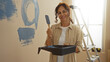 © Krakenimages.com - Middle-aged woman painting wall inside new home while smiling, holding paintbrush and tray, surrounded by ladders and tools, creating cozy atmosphere in living room.