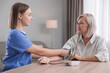 © New Africa - Healthcare worker measuring patient's blood pressure at wooden table indoors