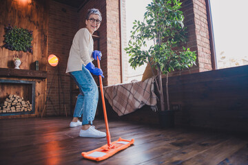 Wall Mural - Charming mature woman joyfully cleaning wooden floor at home in daylight, wearing casual pullover and jeans