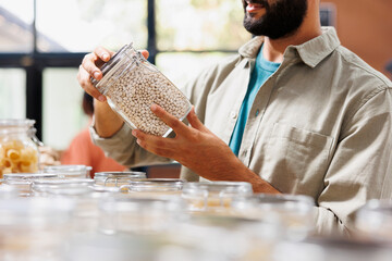 Canvas Print - Close up on glass jar filled with locally grown food product held by male consumer in eco friendly convenience store. Detailed image of person grasping plastic free container having organic beans.