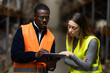 © Nikola Spasenoski - Two professionals wearing safety vests have a focused discussion while reviewing a document in an industrial warehouse setting, showcasing teamwork