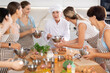© JackF - Elderly woman chef with bowl in hands tells enthusiastic female visitors of culinary courses about unusual traditions of foreign cuisine