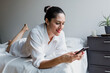 © Marcos - Portrait of mature latin woman using phone and relaxing down on bed laughing at home in Mexico Latin America, hispanic female