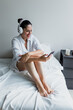 © Marcos - latin adult woman resting on bed using mobile phone and headphones at home in Mexico Latin America, hispanic middle aged female