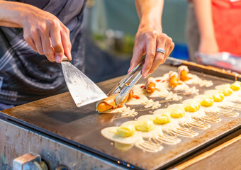 The vendor is rolling a sausage-filled Tokyo pancake on a hot griddle.