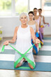 © JackF - Positive fit elderly woman in comfortable sportswear participating in fitness class in well-lit studio, using green elastic resistance band for exercise routine. Healthy lifestyle and pilates concept