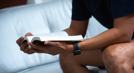 A man is seated comfortably on a sofa, engaged in reading a book. His attire is casual yet neat, such as a shirt and trousers, suggesting a moment of leisure within a serene environment.