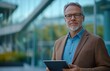 © yevgeniya131988 - Elegant middle-aged businessman with glasses and goatee holding a tablet in front of a modern office