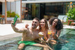 © Dorde - A group of diverse young adults shares a lively moment, capturing a selfie while enjoying refreshing drinks in a sunlit pool.