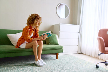 Canvas Print - Young woman reading a book on a green couch in a cozy, modern living room with natural light, reflecting a relaxed and comfortable atmosphere